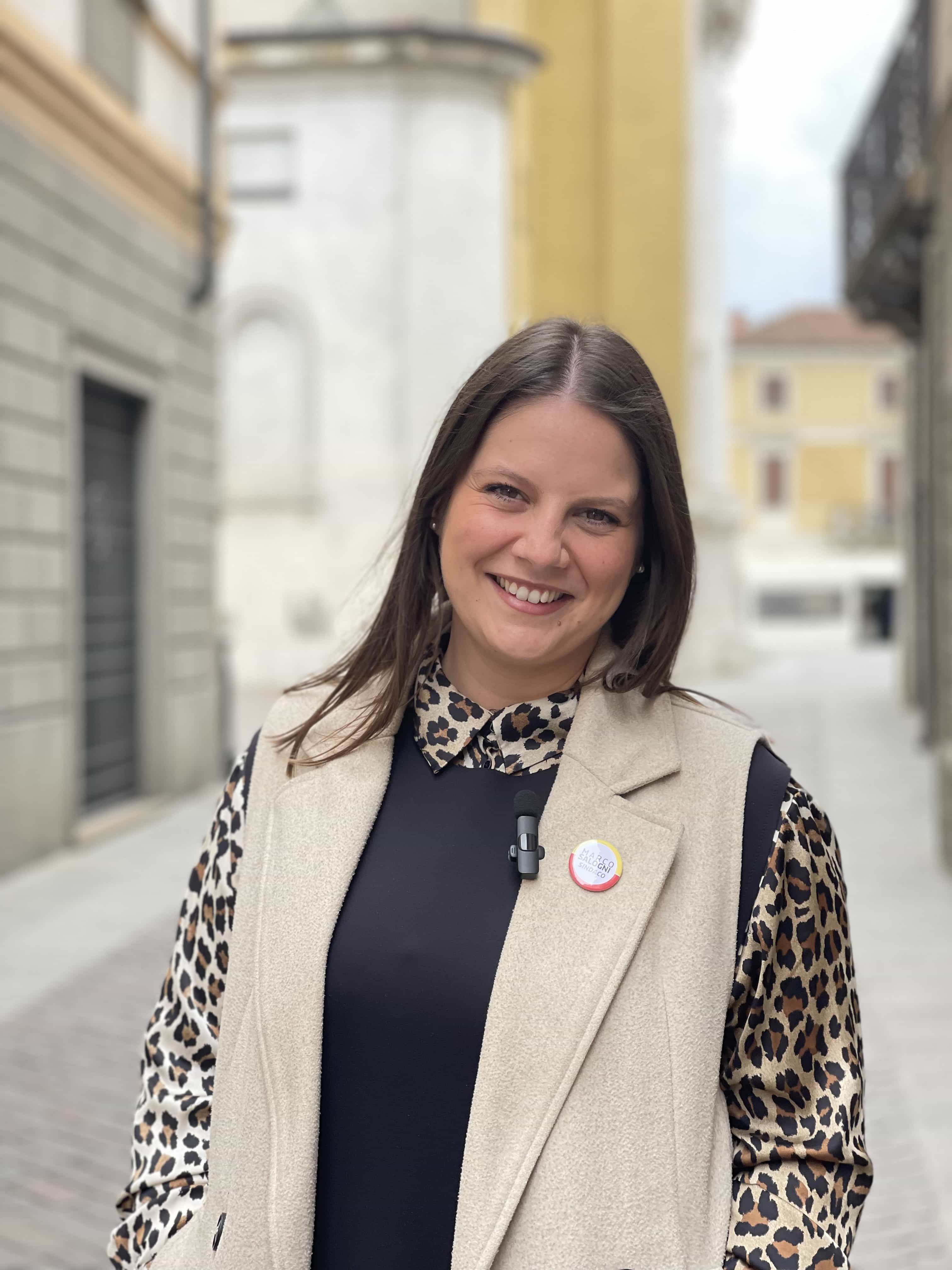 Foto di Camilla a mezzo busto sorridente in giacca smanicata e camicia. Alle spalle via Villatico che imbocca piazza Zanardelli