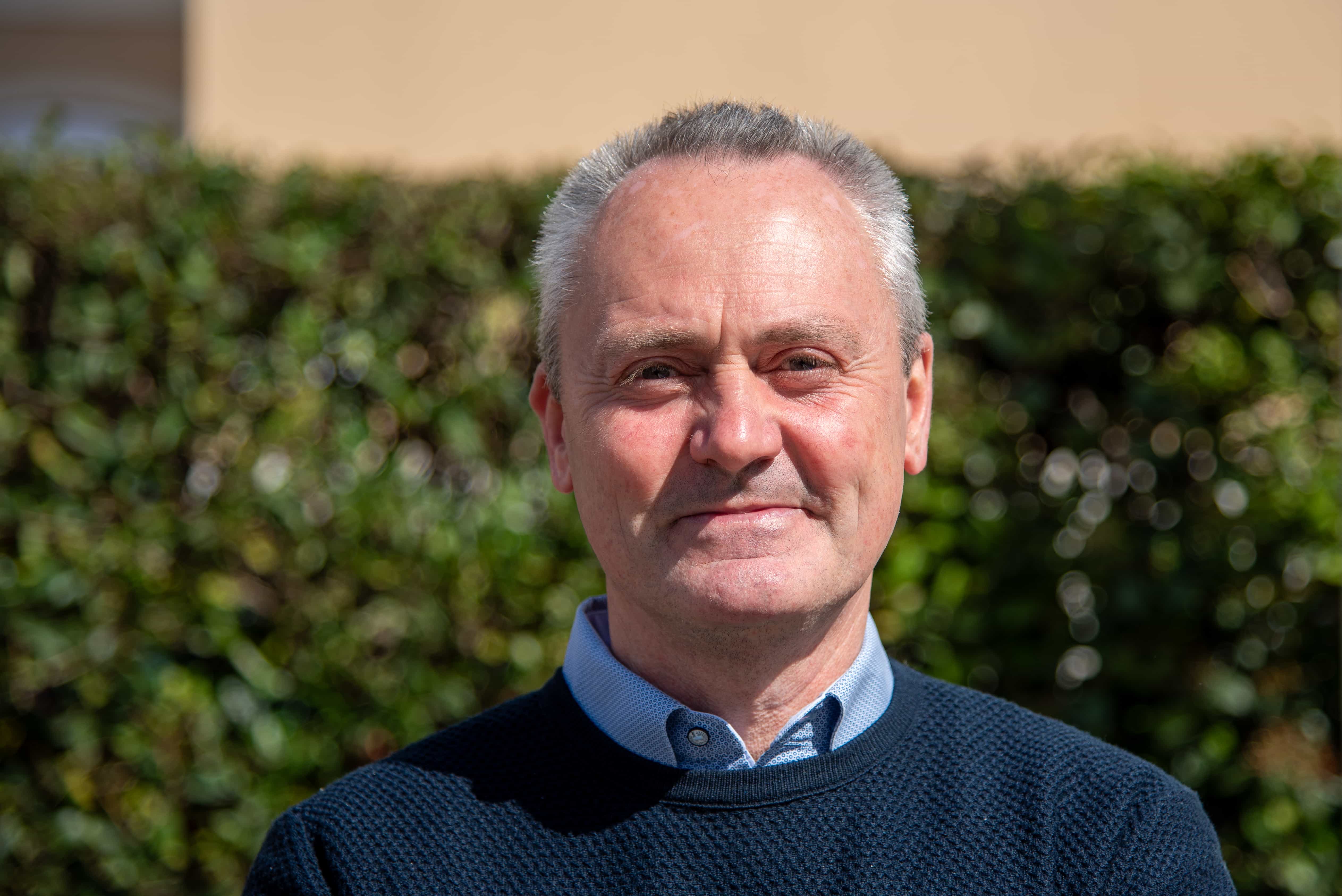 Foto in primo piano di Davide Serlini in camicia e maglione, sorridente con dietro lo sfondo sfocato di una siepe