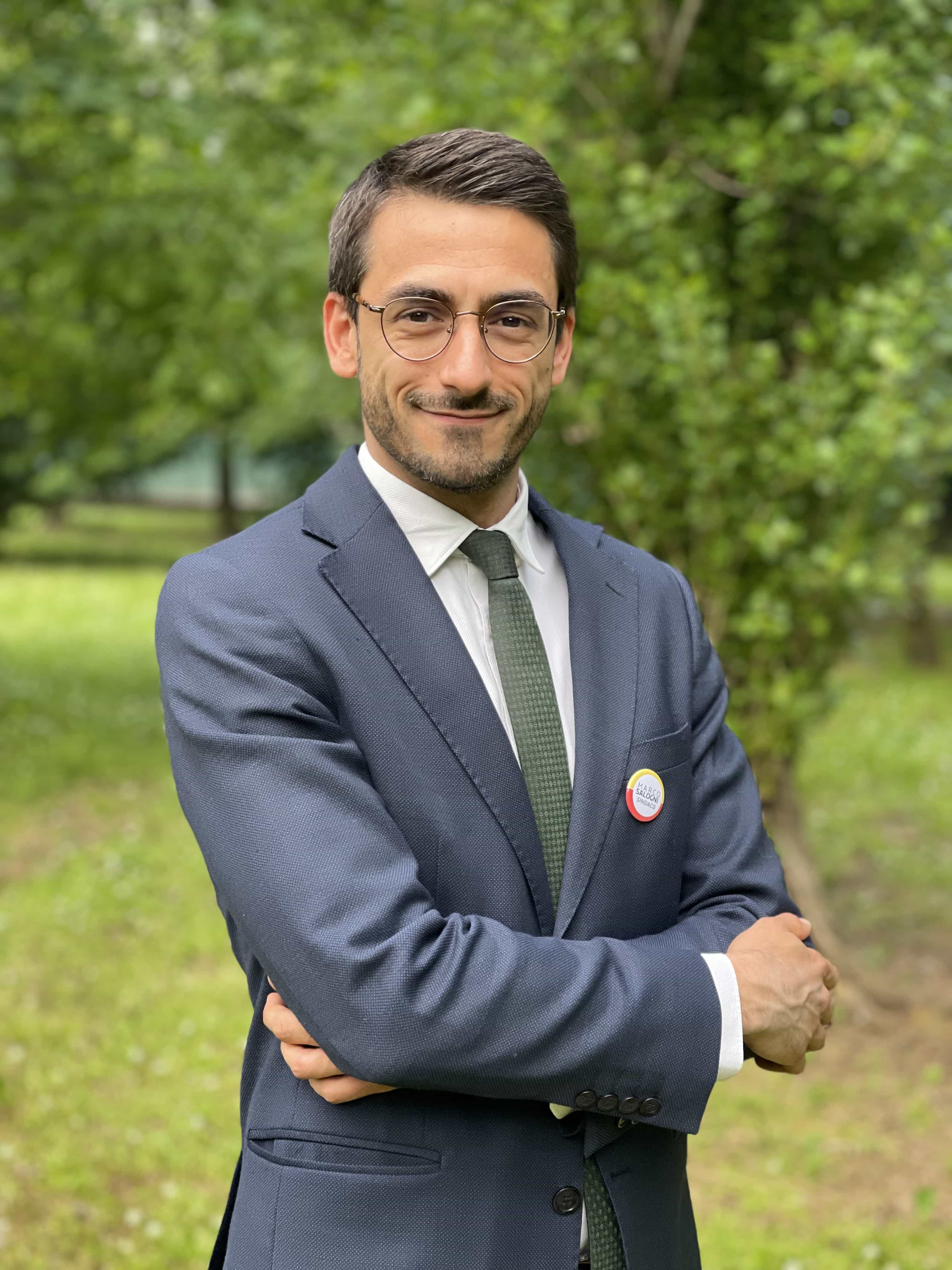 Foto mezzo busto di Davide Adrodegari in giacca, camicia e cravatta e braccia conserte che guarda sorridente l'obiettivo. Alle spalle un parco