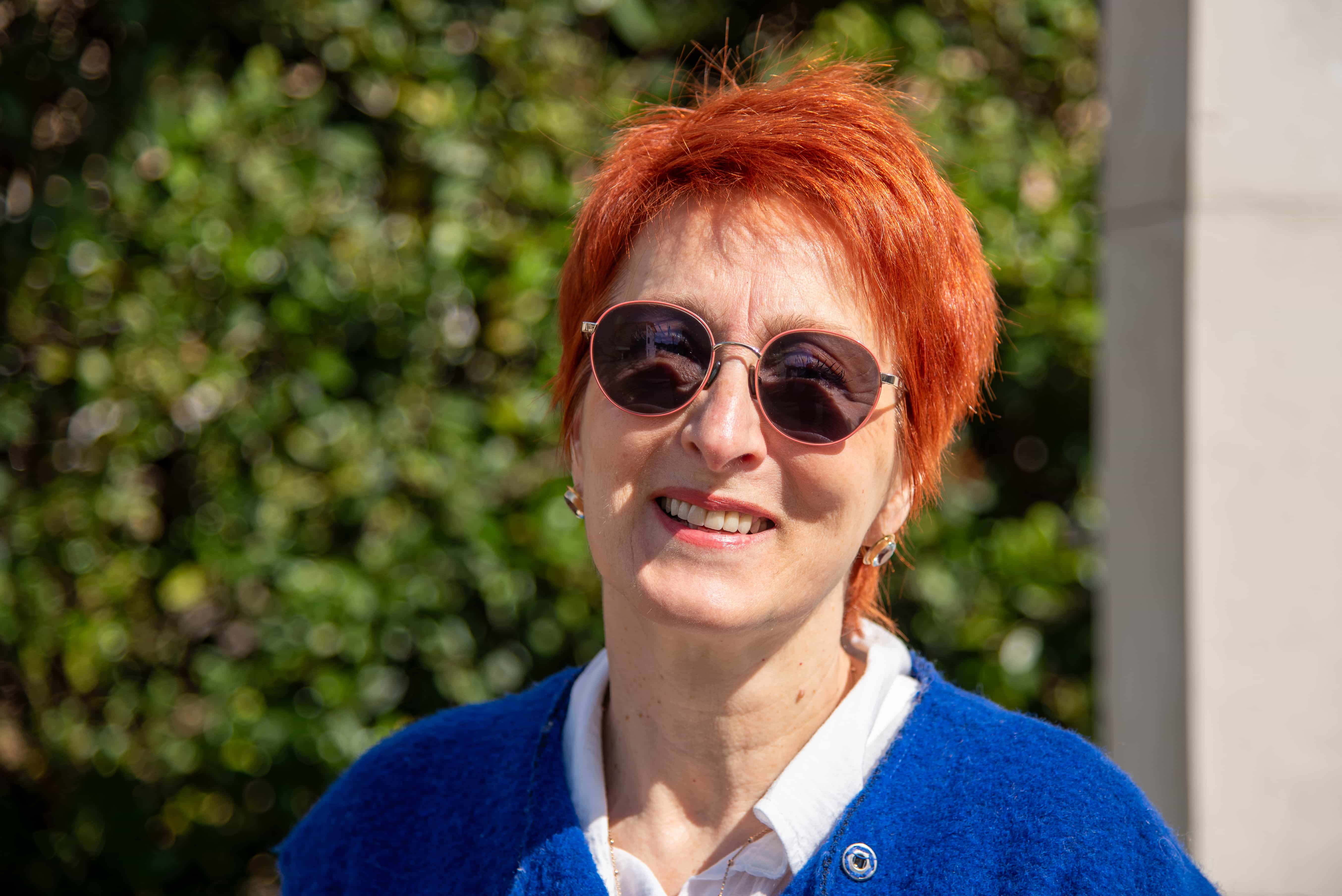 Emanuela Franzoni con maglione blu e camicia che sorride in primo piano. Ha gli occhiali da sole e la foto è molto soleggiata. Alle spalle una siepe 