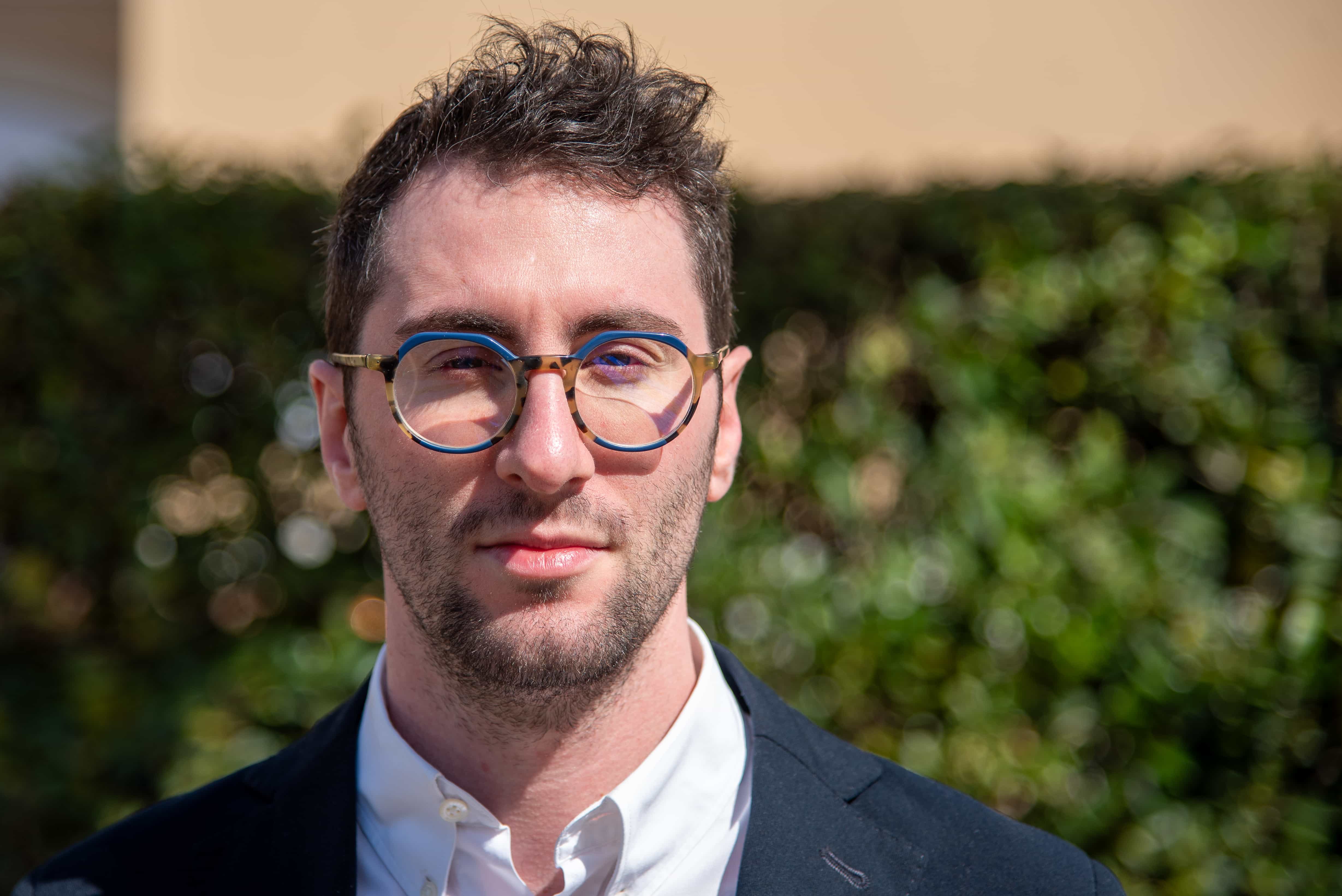 Matteo Ghidinelli in primo piano in camicia col colletto aperto e giacca. Alle spalle, sfocata, una siepe